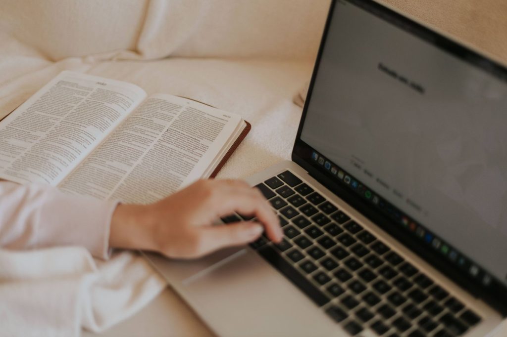 A person multitasks with an open book and a laptop in a cozy indoor setting.