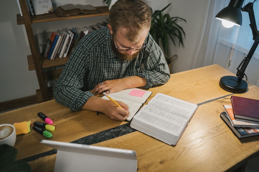 Adult man writing notes and studying at a desk with a Bible and coffee in a cozy room.