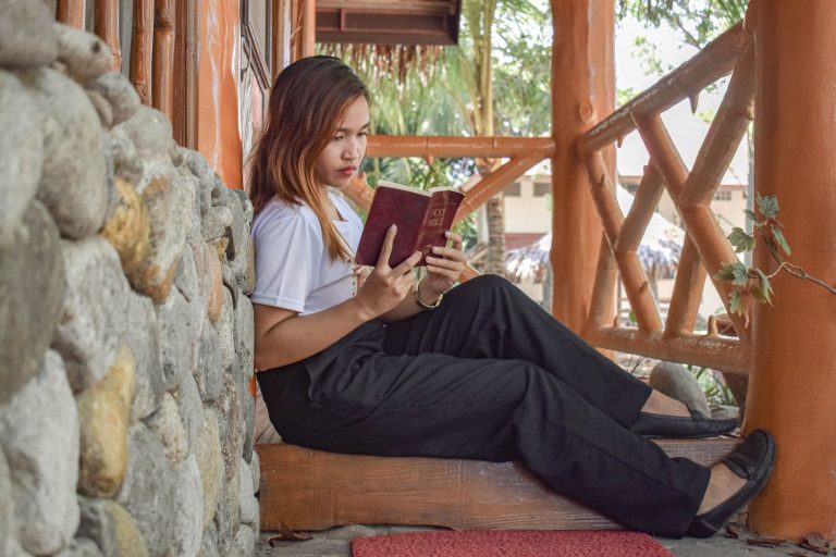 A young woman sitting on a porch reads the Bible, focused and serene in a natural outdoor setting.