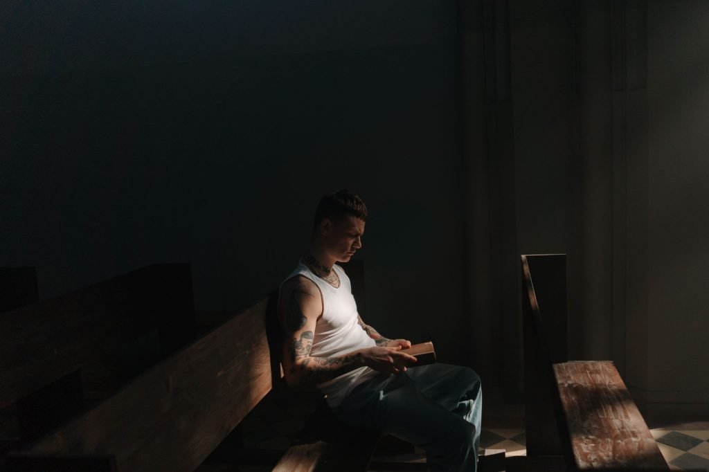 A tattooed man in a white tank top reads a book in dim lighting inside a church.