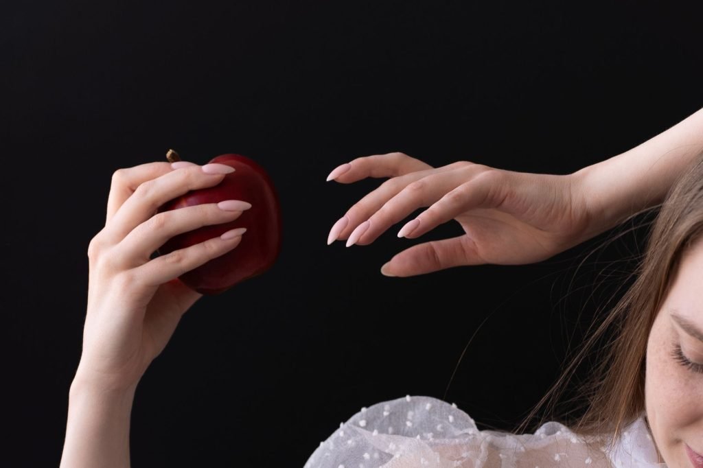Elegant woman's hand holding a red apple against a dark background, symbolizing temptation.
