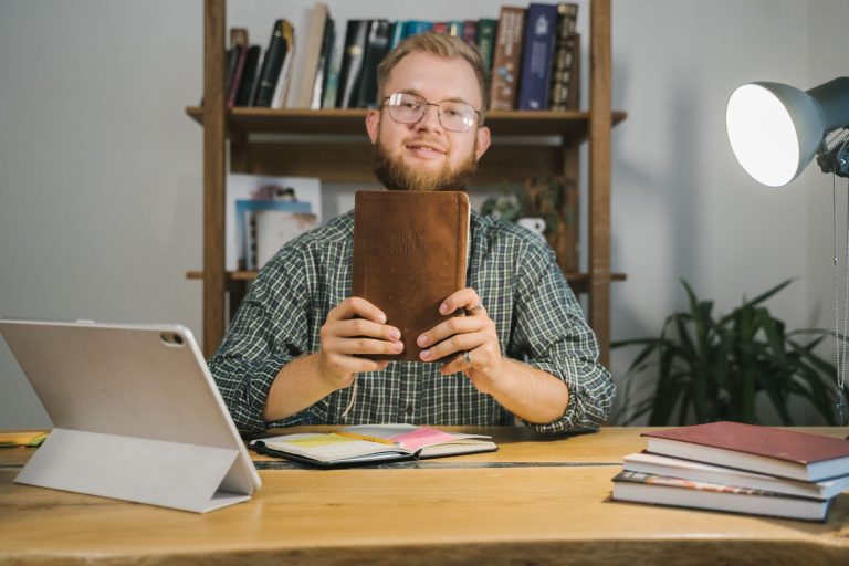 A man in eyeglasses holds a holy Bible at a wooden desk in a study room with shelves of books.