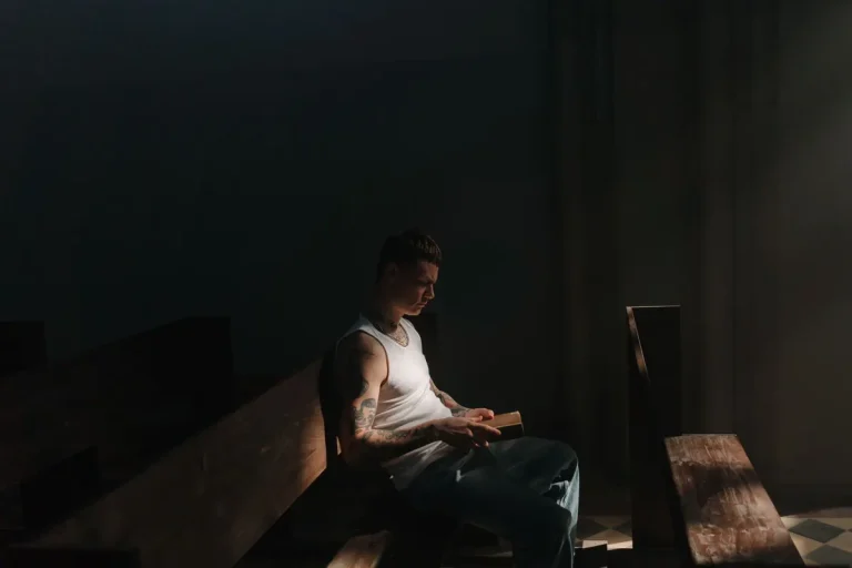 A tattooed man in a white tank top reads a book in dim lighting inside a church.