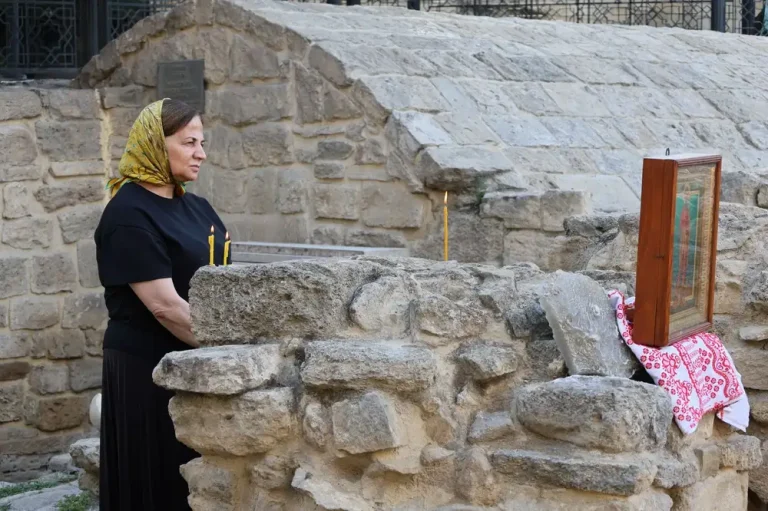 A woman in a headscarf and candles beside religious icon at historic stone ruins. Reflective moment.