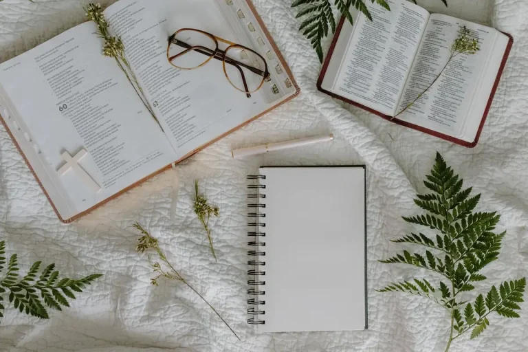 Top view of two open bibles, notebook, glasses, pen, and leaves on a quilted fabric surface.